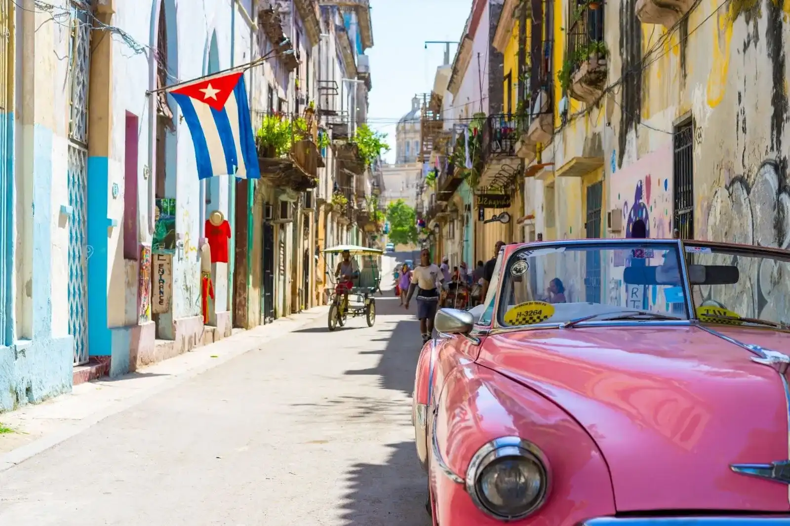 Picture shows a sunny street somewhere in Cuba. The pink cadillac is the right bottom corner. People are walking down the street to the left. 
